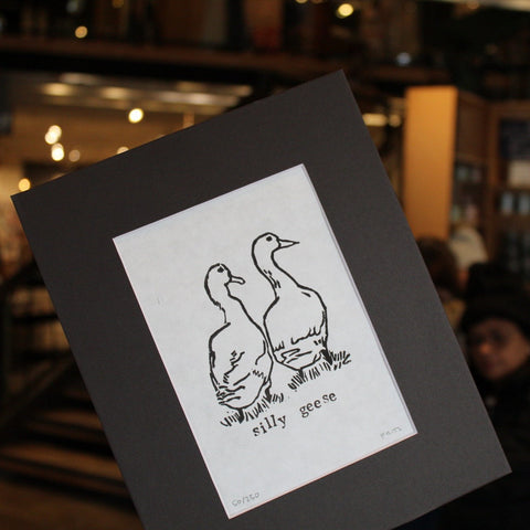 A hand-printed black and white lino print of two geese titled 'Silly geese', presented in a black mat.