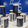 A set of ceramic mugs and a pitcher with a navy blue and white vertical stripe pattern, arranged on a surface in front of a blue sign that reads 'Snacks'.