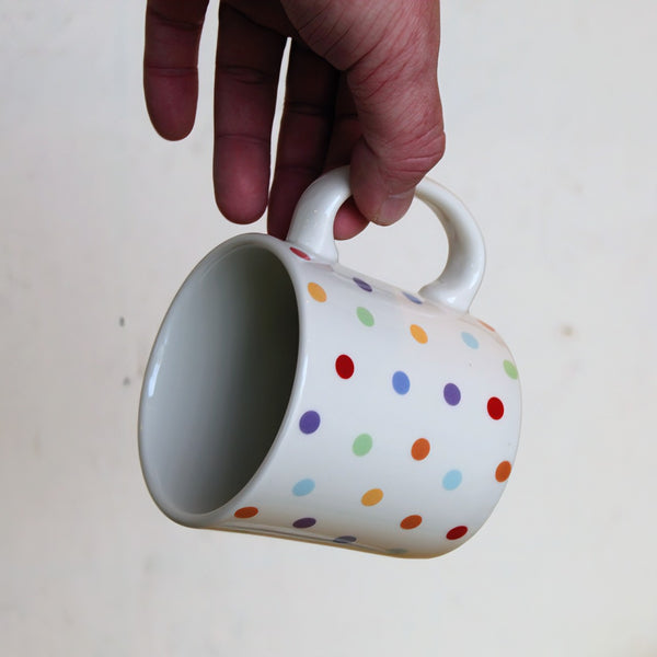 Hand holding a white mug with colorful polka dots on a light background