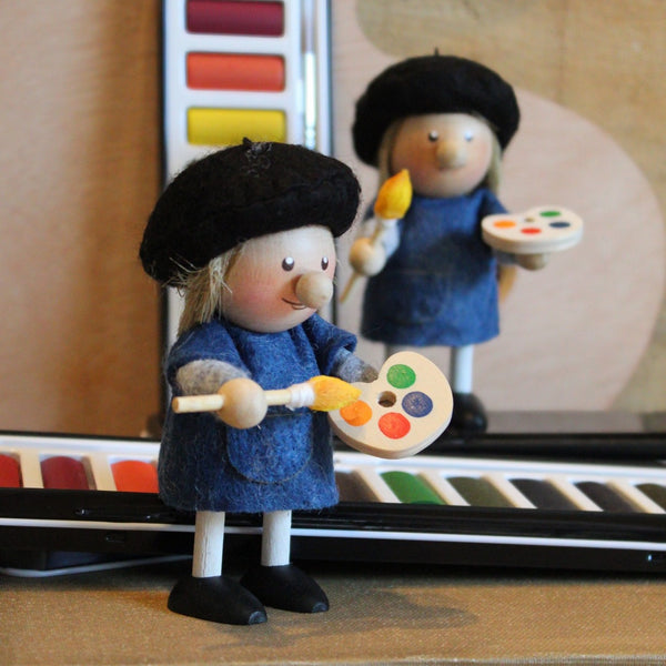 A small handmade artist, wearing a wool felt black beret and blue apron, holding a paintbrush and a wooden paint palette.  with paint palettes in the background.