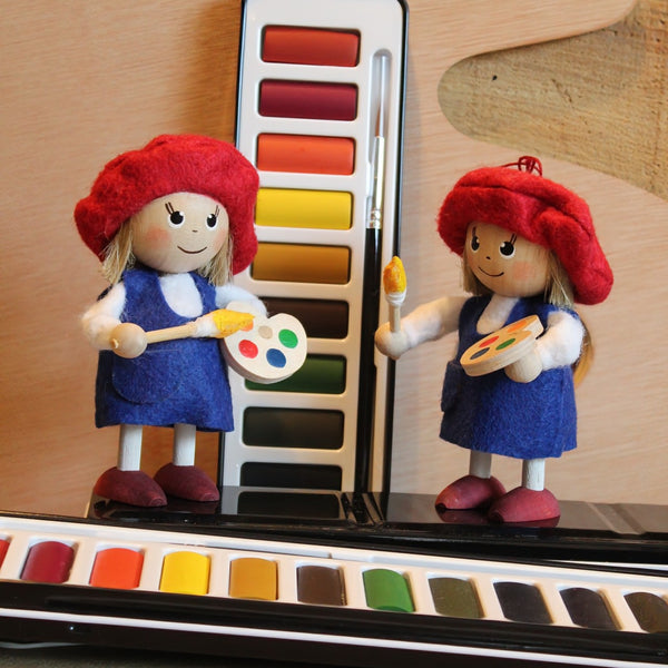 A small handmade wooden artist wearing a red beret and blue painter's apron, holding a paintbrush and a paint palette, posed in front of a watercolor paint palette.