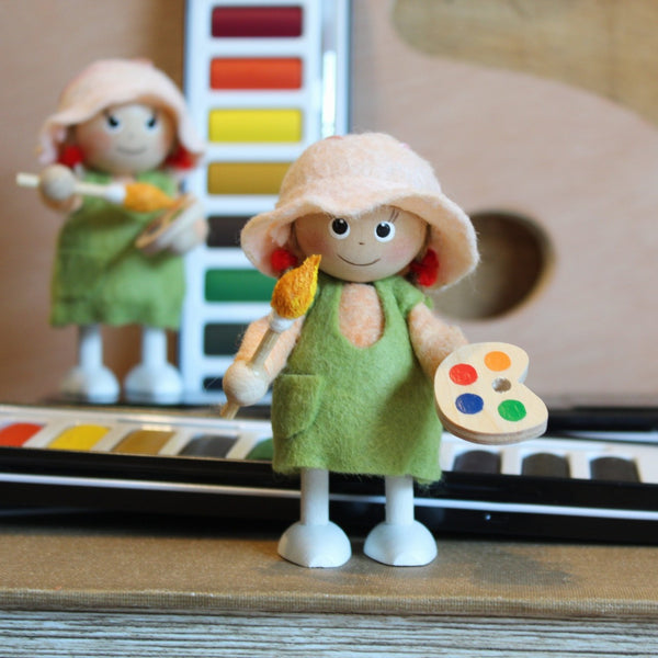 A small handmade artist stands in front of a colorful watercolor palette, holding a paintbrush and a wooden paint palette with multicolored paints. A second similar doll and palette are blurred in the background.
