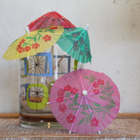 A retro glass with a selection of The Original Cocktail Umbrellas in and around it on a white background. 