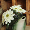 close up image of a green stonewate watering pitcher in the shape of a frog with an open mouth being used as a vase - holding white flowers on a wooden surface