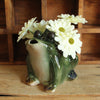 Green stonewate watering pitcher in the shape of a frog with an open mouth being used as a vase - holding white flowers on a wooden surface