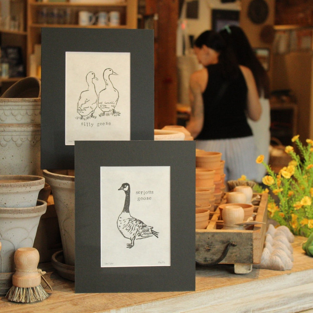 Two art prints featuring goose illustrations displayed on a rustic wooden table in a pottery and craft shop, with a woman blurred in the background.