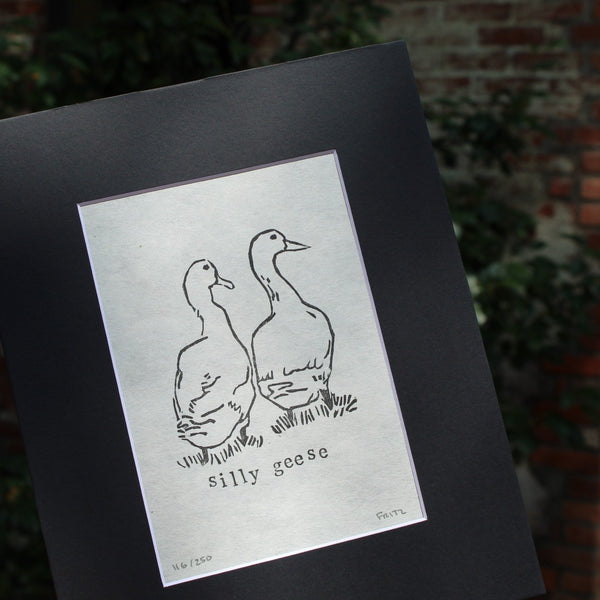 A black and white art print depicting two silly geese, mounted on a wide black mat board. The print is photographed at an angle against a blurred outdoor background of green foliage and a brick wall.