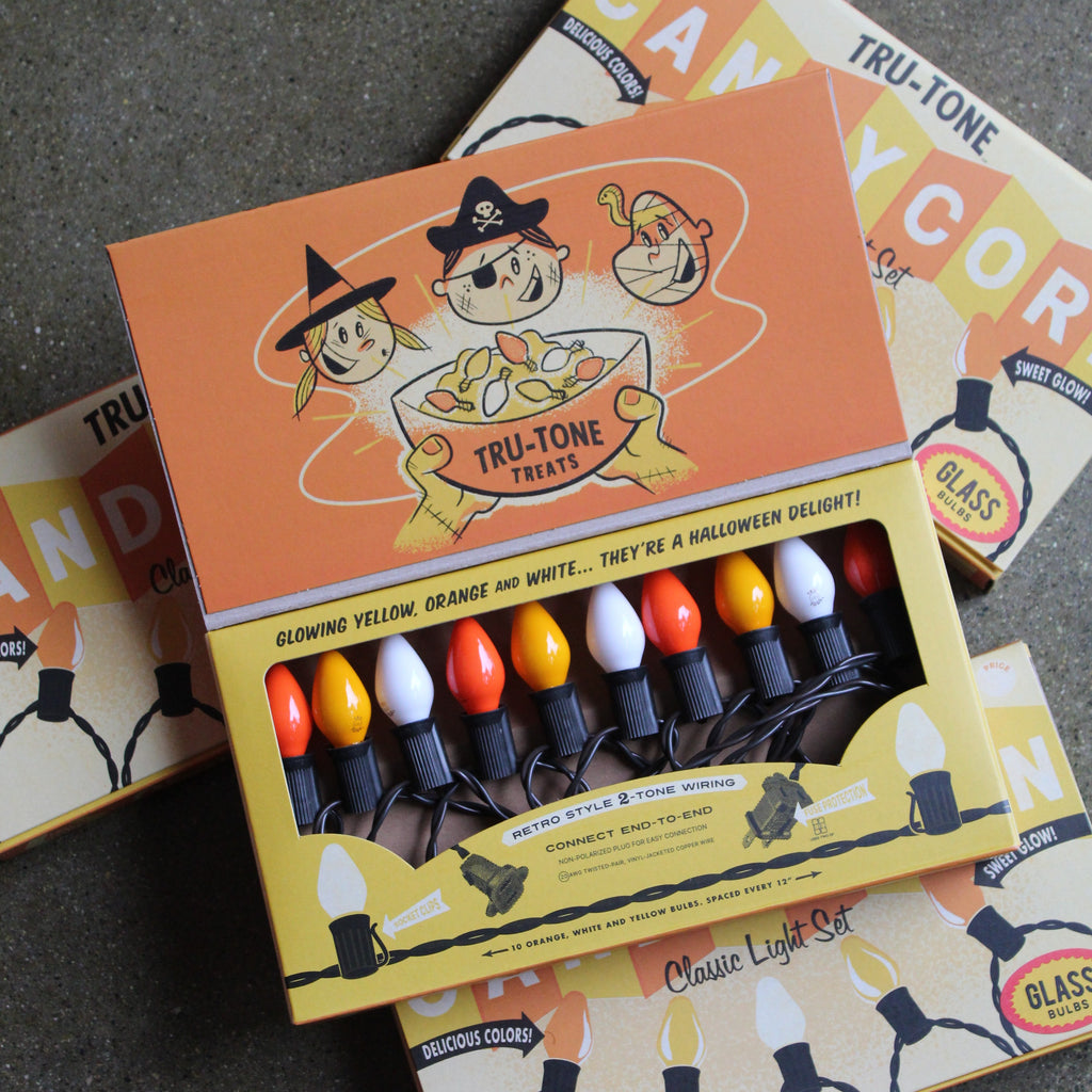 A boxed set of retro-style Halloween string lights displayed open on top of matching product boxes. The lights have glowing yellow, orange, white bulbs on a black cord, the packaging features orange and yellow artwork with cartoon characters and the text 'TRU-TONE TREATS' and 'Classic Light Set'. 