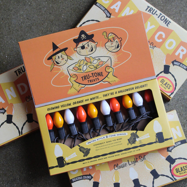A boxed set of retro-style Halloween string lights displayed open on top of matching product boxes. The lights have glowing yellow, orange, white bulbs on a black cord, the packaging features orange and yellow artwork with cartoon characters and the text 'TRU-TONE TREATS' and 'Classic Light Set'. 