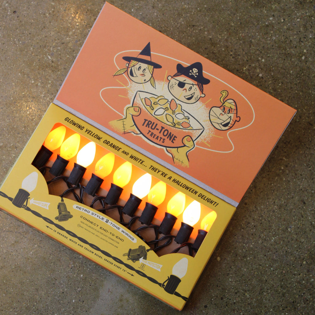 A package of Halloween 'Tru-Tone Treats' string lights, shown open on a concrete floor. The box is orange and yellow with cartoon illustrations of a witch, a ghost, and a pumpkin.