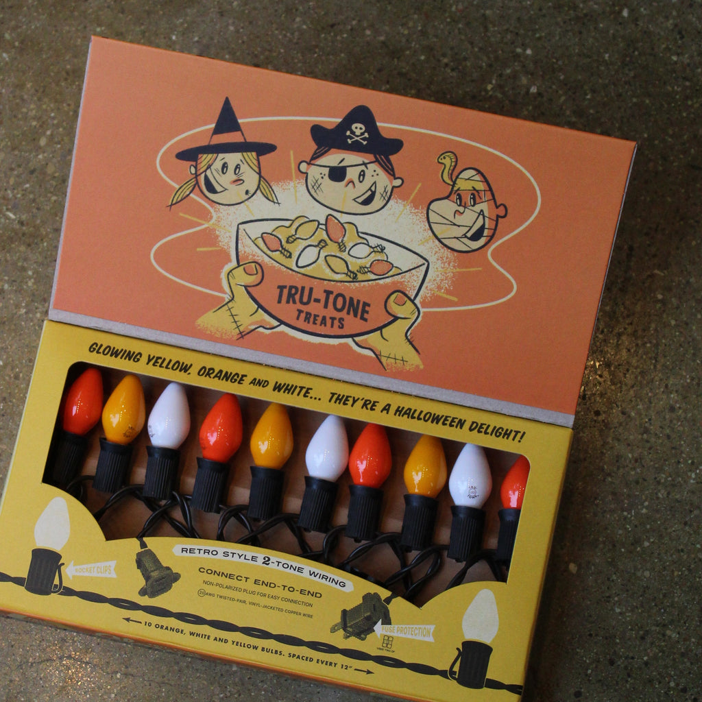 A box of Tru-Tone Halloween string lights with retro-style bulbs in orange, yellow, white, on a black cord, shown in its illustrated packaging on a dark textured surface.