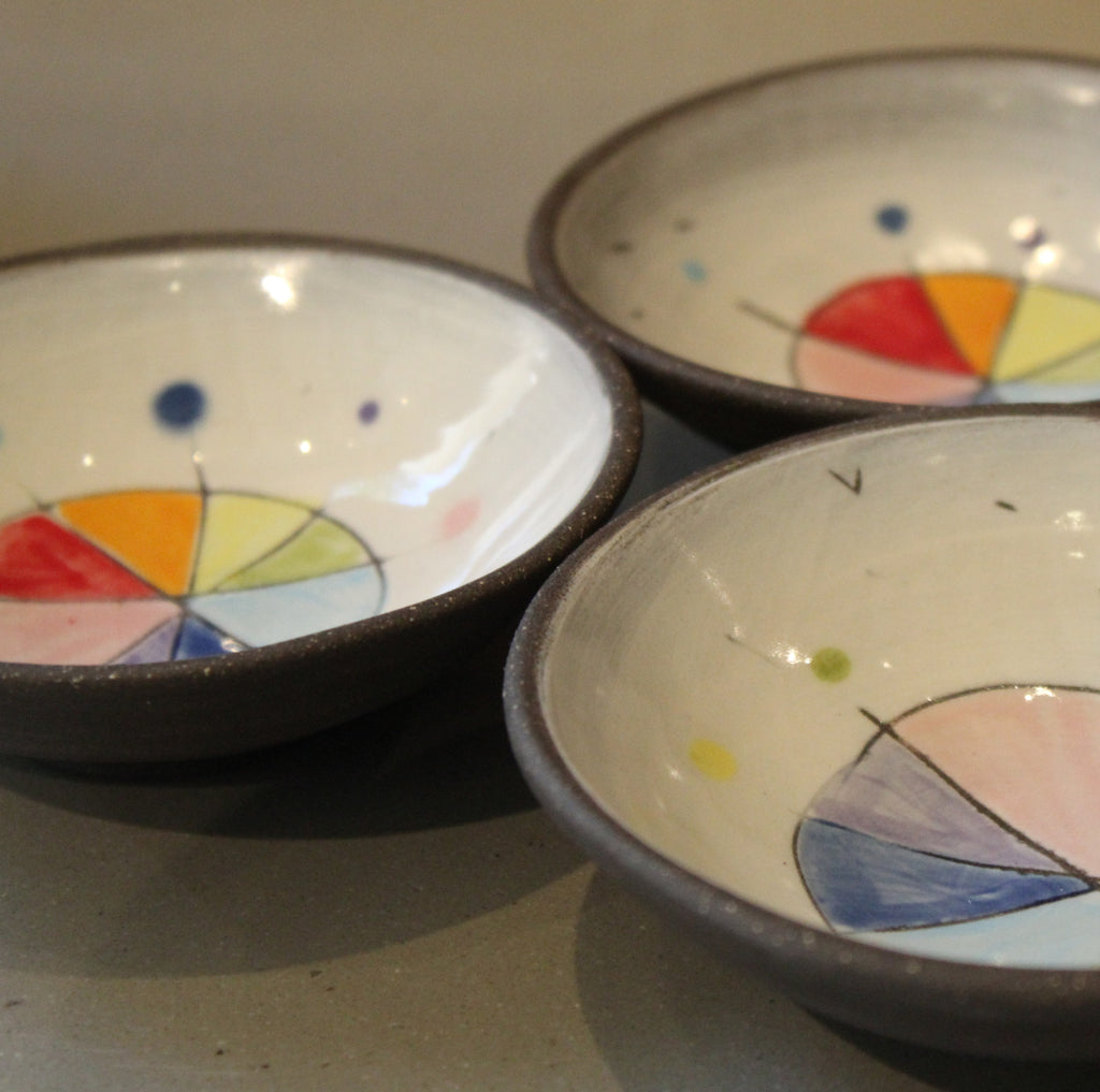 Three handmade ceramic bowls with dark brown rims and colorful painted geometric designs inside, arranged on a neutral gray surface.