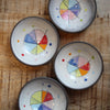 Four small white ceramic bowls with dark rims, decorated with colorful painted color wheels and dots, arranged on a rustic wooden surface.
