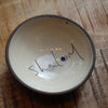 A handmade ceramic bowl with a dark brown rim and a cream-colored interior, featuring the word 'Shalom' written in black script with a small blue dot accent, placed on a wooden surface.