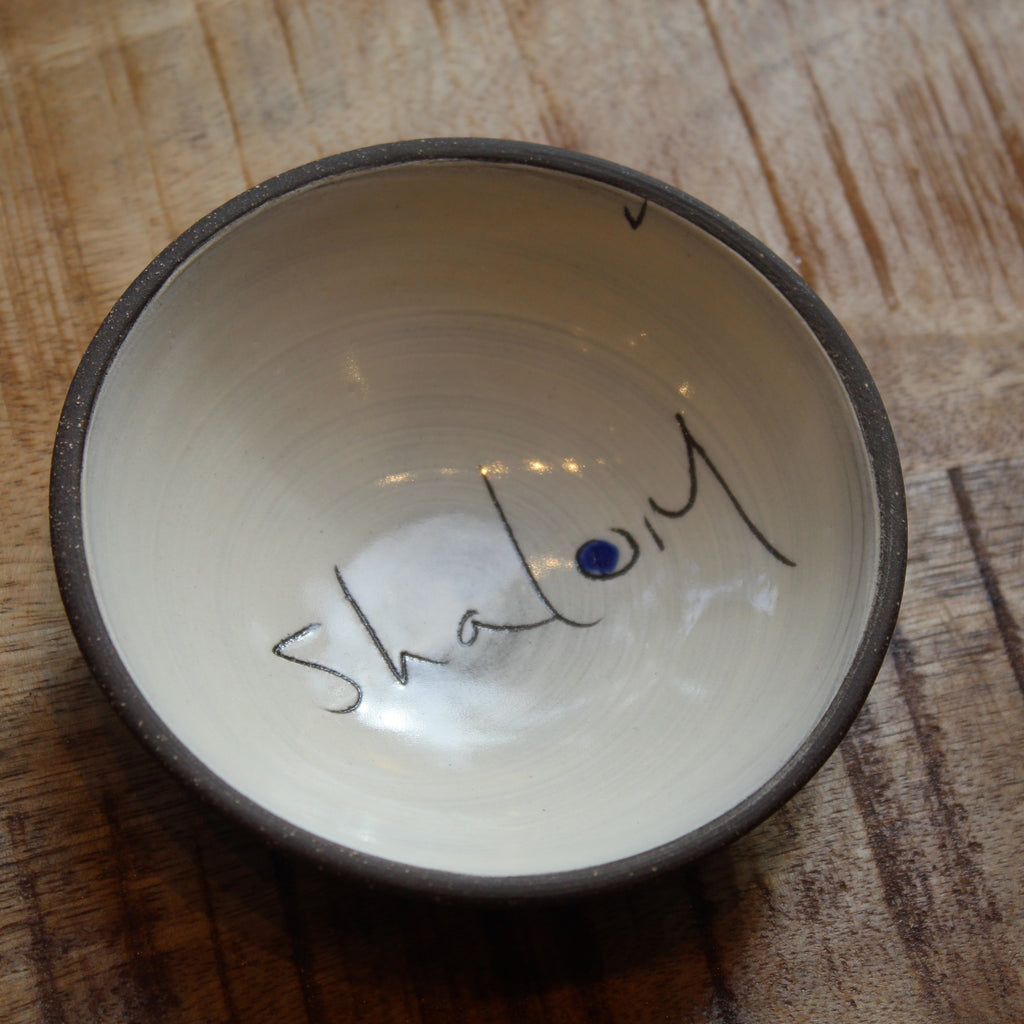 A handmade ceramic bowl with a dark brown rim and a cream-colored interior, featuring the word 'Shalom' written in black script with a small blue dot accent, placed on a wooden surface.
