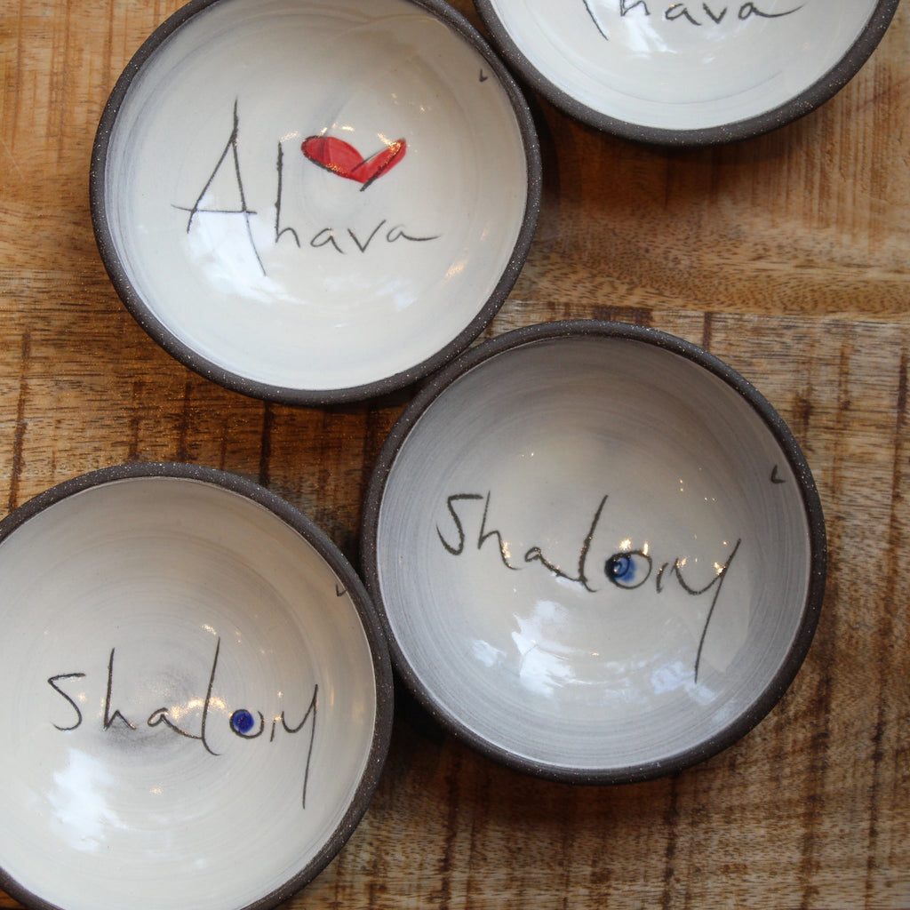 Four handmade ceramic bowls with dark rims, arranged on a wooden surface. Each bowl is decorated with a different hand-painted word or symbol inside: 'Ahava' with a red heart, 'Shalom' with a blue dot.