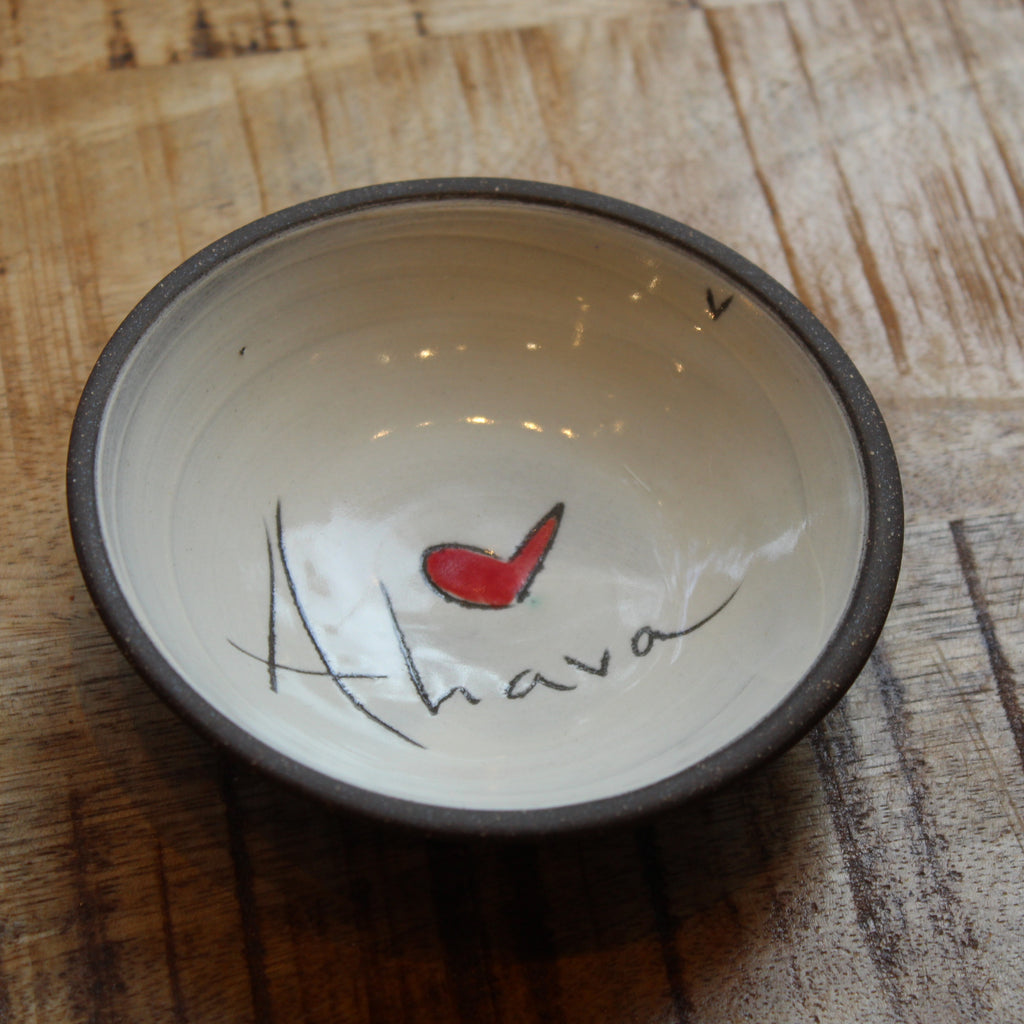 A handmade ceramic bowl with a dark brown rim and a cream-colored interior, featuring a hand-painted red heart and the word 'Ahava' in black script on the inside. The bowl is placed on a rustic wooden surface.