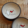 Two handmade ceramic bowls with dark gray rims and cream interiors, decorated with a red heart and the word 'Ahava' in black script, displayed on a wooden surface.