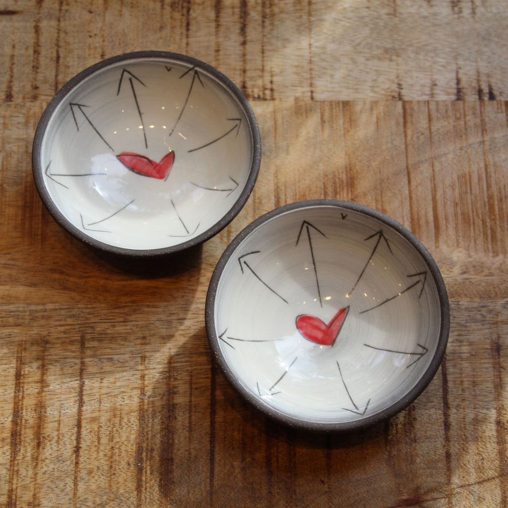 Two small round ceramic bowls with dark rims, featuring a white interior decorated with black radial arrows and a small red heart near the bottom of each bowl, set on a wooden surface.