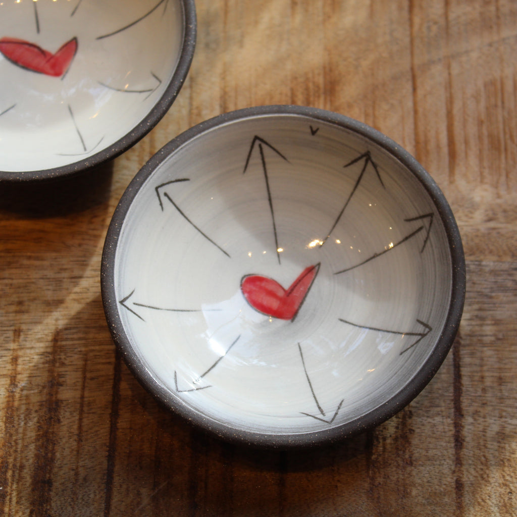 A handmade ceramic bowl with a dark rim, featuring a white interior decorated with black hand-drawn arrows radiating from a small red heart, shown on a wooden table.