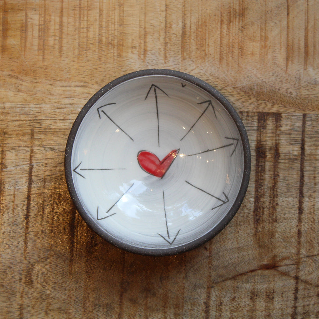 A small round ceramic bowl with a dark rim, featuring a white interior decorated with black radial arrows and a small red heart in the center, placed on a wooden surface.