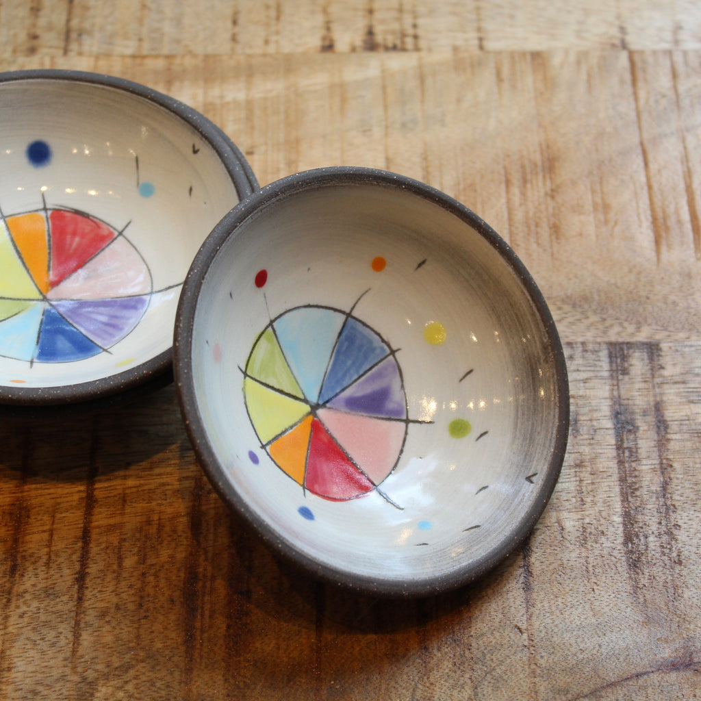 Two handmade ceramic bowls with dark rims and colorful painted geometric designs inside, displayed on a rustic wooden surface.