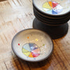 A small handmade ceramic bowl with a colorful geometric pattern inside, shown on a wooden table with a stack of similar bowls behind it.