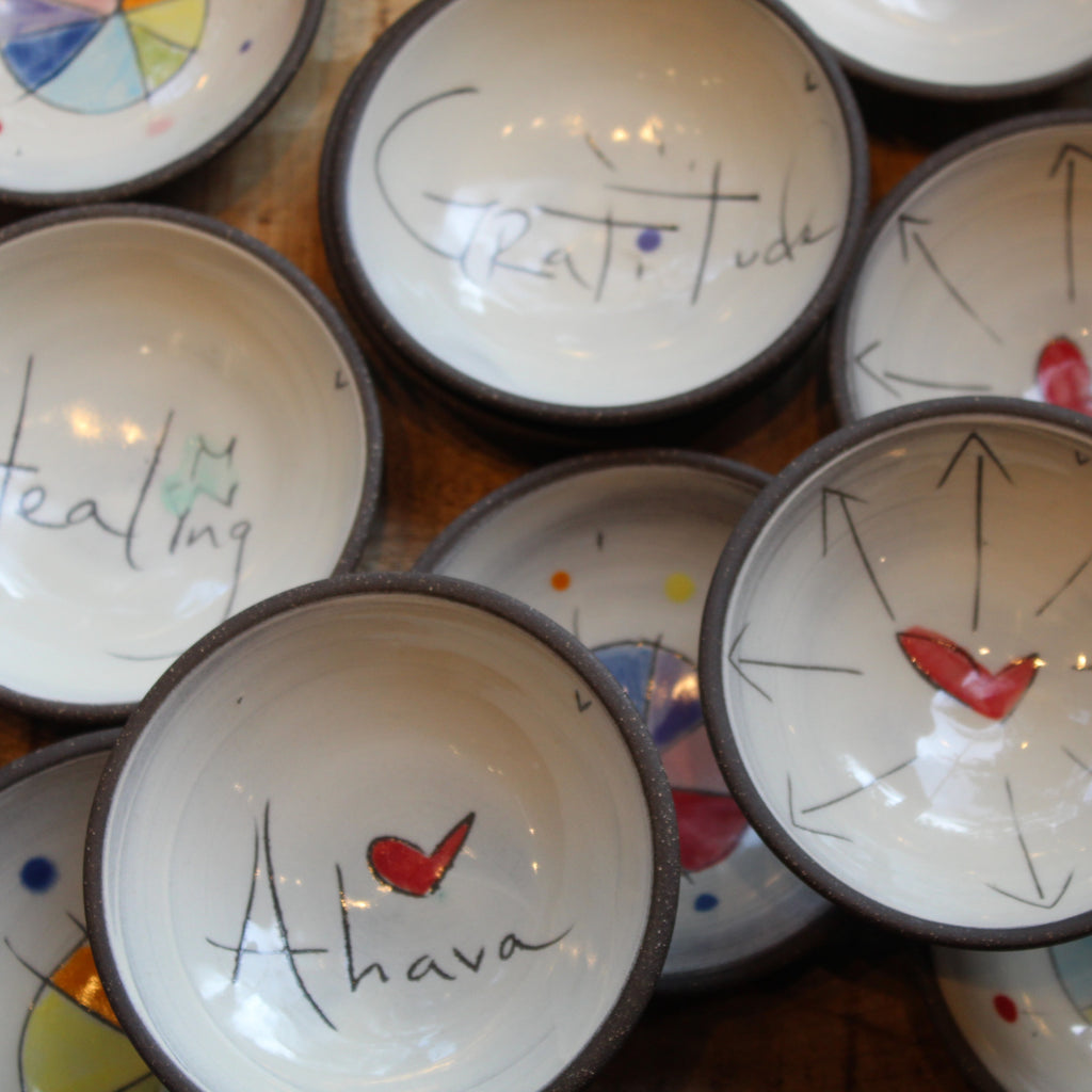 A close-up view of multiple handmade ceramic bowls with dark rims, each decorated with a different inspirational word or phrase and small colorful painted details, including red hearts and blue, yellow, and green accents.