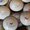 A close-up view of multiple handmade ceramic bowls with dark rims, each decorated with a different inspirational word or phrase and small colorful painted details, including red hearts and blue, yellow, and green accents.
