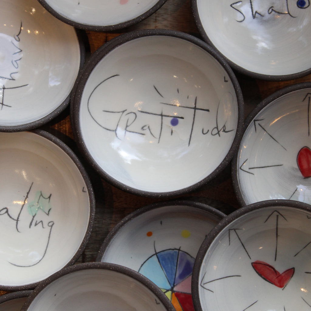 A close-up arrangement of small white ceramic bowls with dark rims, each decorated with hand-painted symbols, arrows, and words including 'Gratitude' on the central bowl.