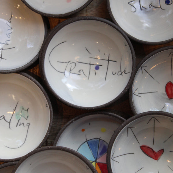A close-up arrangement of small white ceramic bowls with dark rims, each decorated with hand-painted symbols, arrows, and words including 'Gratitude' on the central bowl.