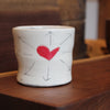 A small white ceramic cup with a hand-painted red heart in the center, surrounded by simple black arrow lines, resting on a wooden surface.