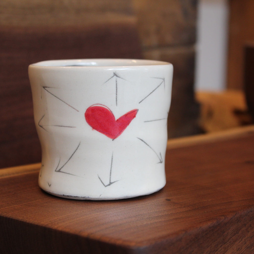 A small white ceramic cup with a hand-painted red heart in the center, surrounded by simple black arrow lines, resting on a wooden surface.