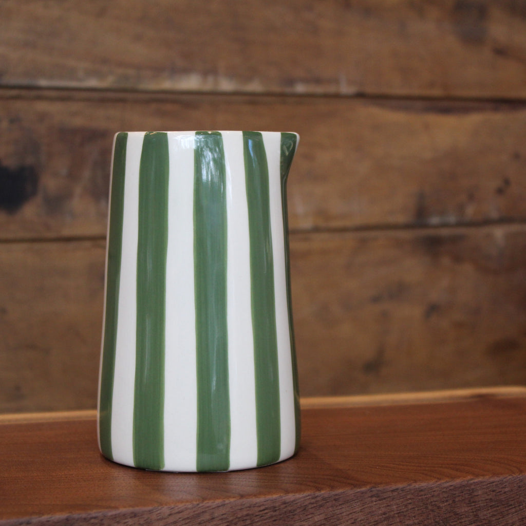 A glossy ceramic pitcher with vertical green and white stripes, standing on a wooden surface against a rustic wooden plank background.