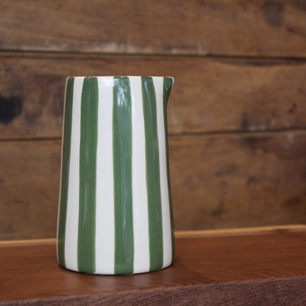 A glossy ceramic pitcher with vertical green and white stripes, standing on a wooden surface against a rustic wooden plank background.
