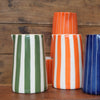 A set of ceramic pitchers with vertical stripes, including green and white, orange and white, and blue and white, arranged on a wooden surface against a rustic wood plank background.