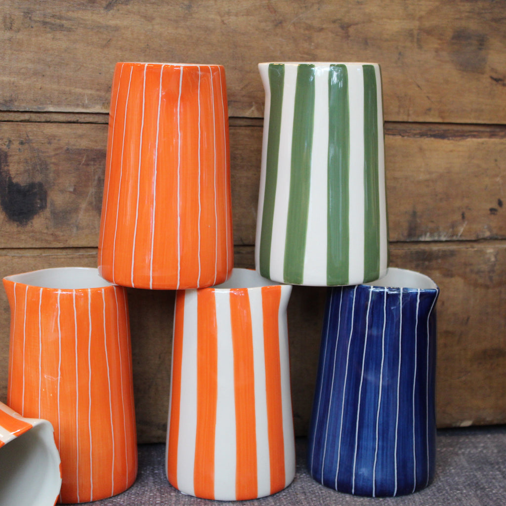A set of five handmade ceramic pitchers with vertical stripes, stacked together against a rustic wooden background. The pitchers feature color combinations of orange, green, blue, and orange/white stripes.