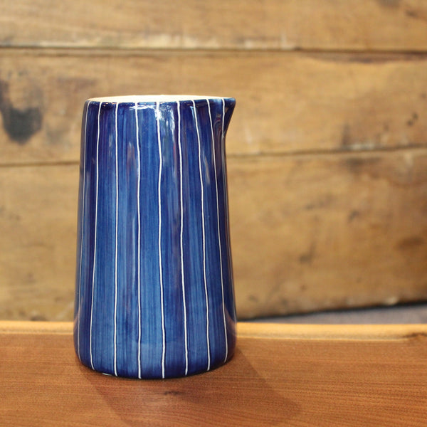 A blue ceramic pitcher with vertical white stripes, standing on a wooden surface against a rustic wooden plank background.