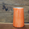 An orange ceramic pitcher with vertical white stripes, standing on a wooden surface against a rustic wooden plank background.