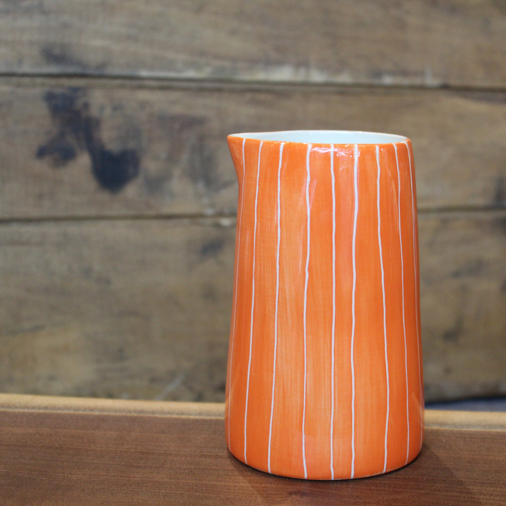An orange ceramic pitcher with vertical white stripes, standing on a wooden surface against a rustic wooden plank background.