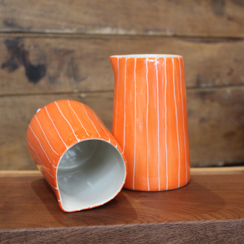 An orange ceramic pitcher with white vertical stripes, shown standing and lying on its side on a wooden surface against a rustic wood plank background.
