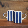 Two ceramic mugs with vertical blue stripes on a white glaze, placed on a wooden surface against a rustic wood plank background.