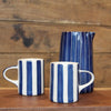 Two ceramic mugs with a blue and white vertical stripe pattern, two smaller mugs in the foreground and one larger pitcher in the background, set on a wooden surface against a rustic wooden wall.