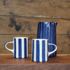 Two white ceramic mugs with blue vertical stripes and a matching tall pitcher, displayed on a wooden surface against a rustic wooden plank background.