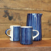 Two ceramic mugs and a matching pitcher in a blue and white striped glaze, arranged on a wooden surface against a rustic wood plank background.