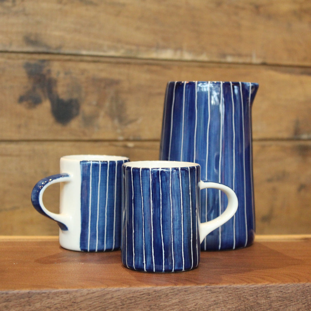 Two ceramic mugs and a matching pitcher in a blue and white striped glaze, arranged on a wooden surface against a rustic wood plank background.