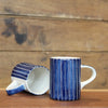 Two ceramic mugs with a blue and white vertical stripe pattern, one standing upright and the other lying on its side to show the interior, set against a rustic wooden background.